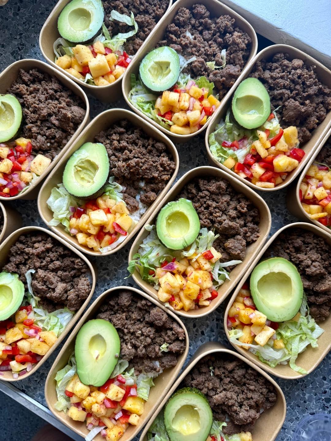 Meal prep containers with seasoned ground beef, lettuce, pineapple salsa, and avocado, made by Andy Bob's